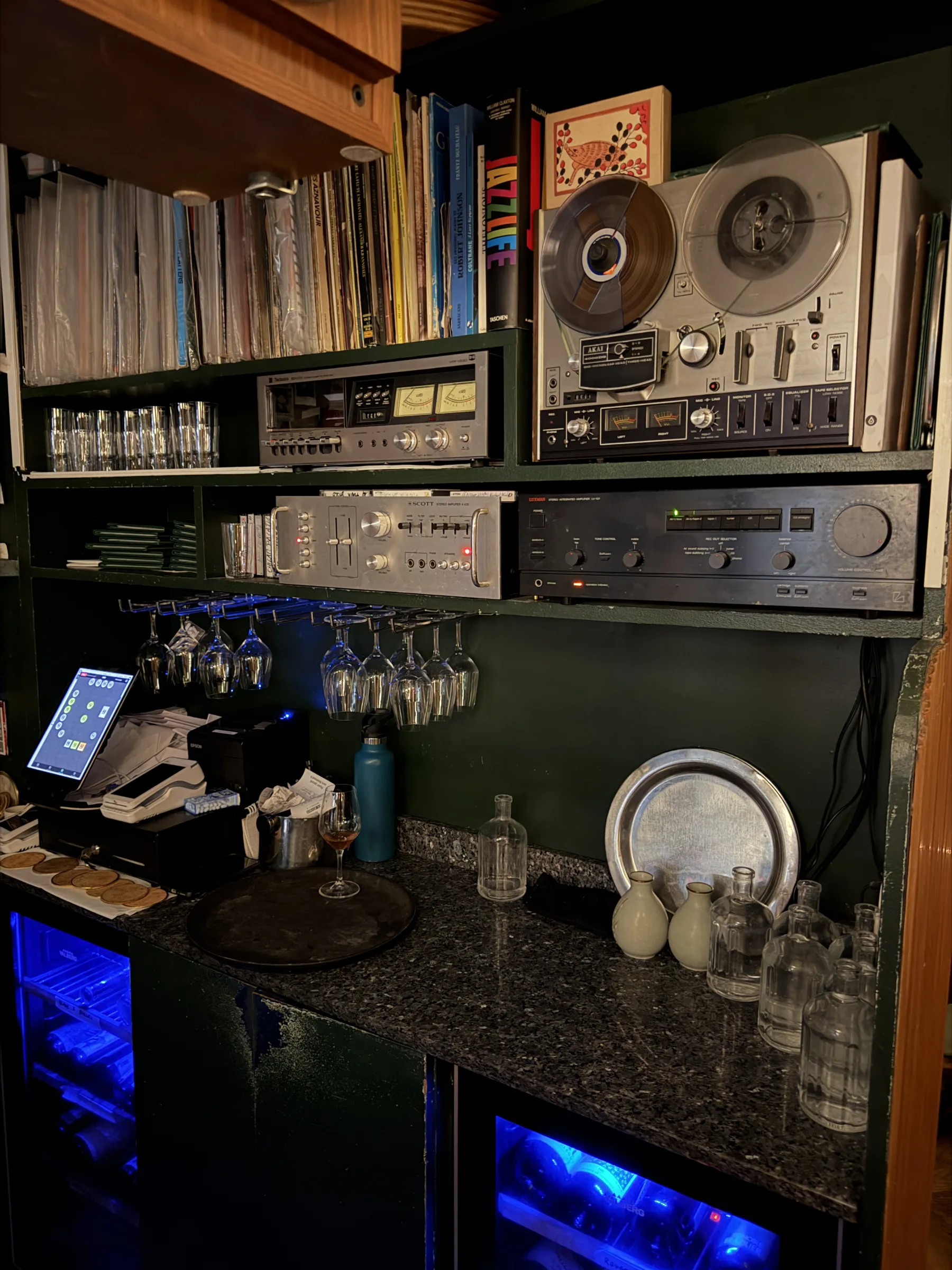 A reel-to-reel tape machine above a rack of amplifiers and a wall of records at Mesures, Paris